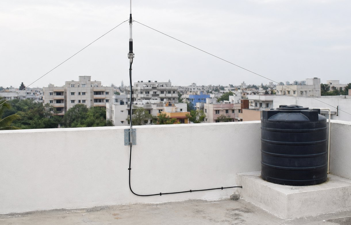 A flat concrete roof with a low parapet. A guyed vertical aerial is bolted to the parapet with a metal bracket; three guy wires run down to anchors. Coaxial cable leaves the feedpoint, runs down the wall and along the roof under cable clips, past a black plastic water tank on a raised plinth. Low-rise apartment blocks and palm trees fill the background.